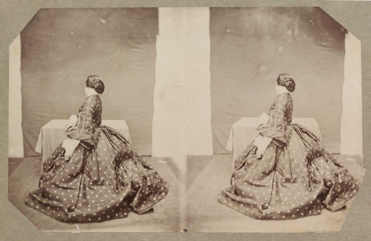 A vintage sepia-toned stereoscopic photograph showing a woman in a polka dot dress seated and looking away from the camera, with her image doubled side by side. The background is minimalistic, with just a draped table, suggesting a studio setting. The edges of the photograph are cut in an octagonal shape.