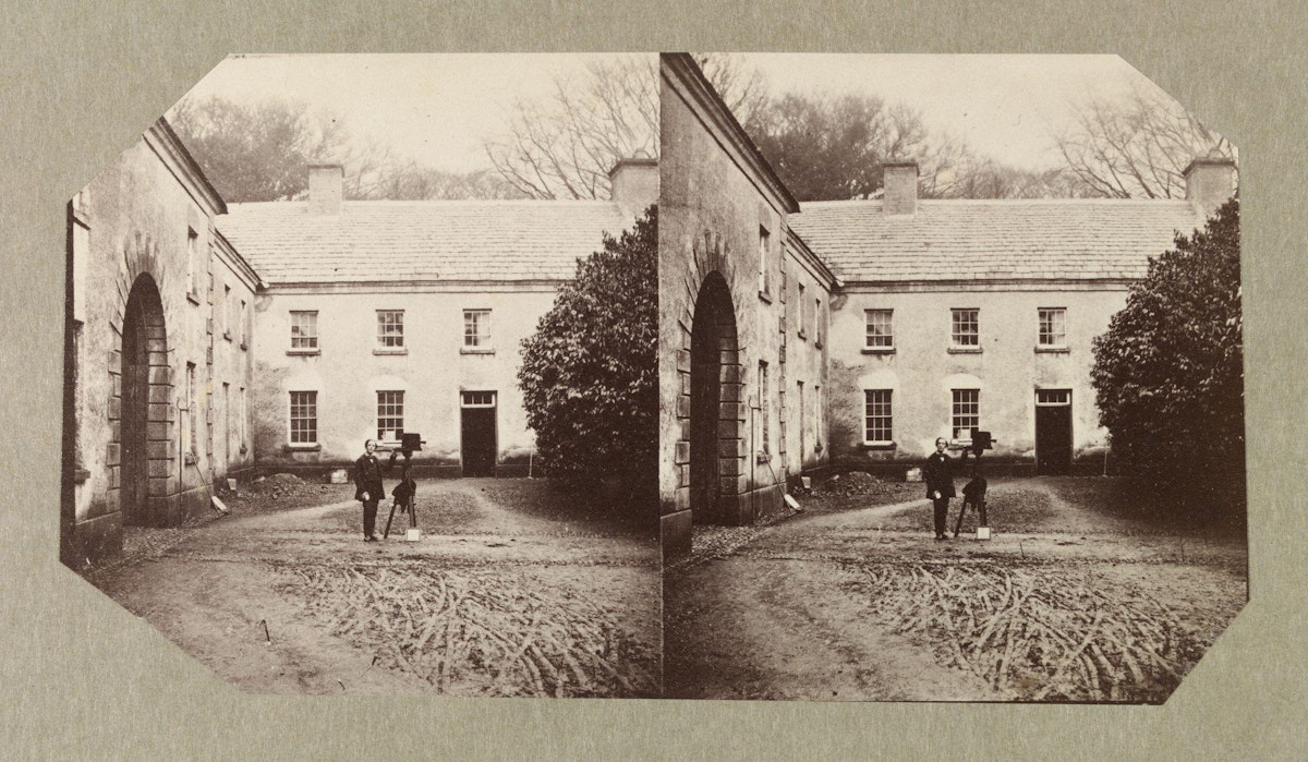 A sepia-toned stereoscopic photograph depicting two individuals standing in front of a two-story country house with an arched entrance, captured in a rural setting. The image is doubled side by side for a 3D effect when viewed through a stereoscope, and the photograph has octagonal cut edges.
