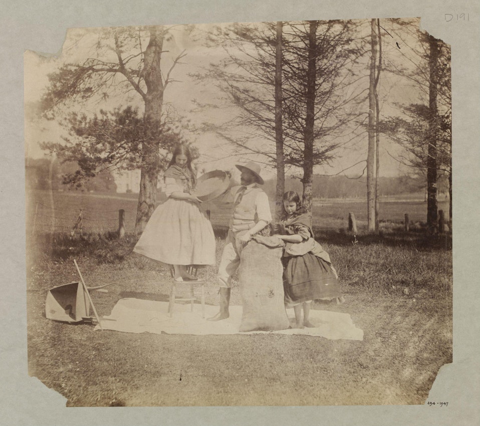 A sepia-toned vintage photograph showing two girls and a boy in a rural setting. One girl is standing on a chair, the boy is seated with a hat, and the other girl is standing beside him, with a fallen parasol on the ground.