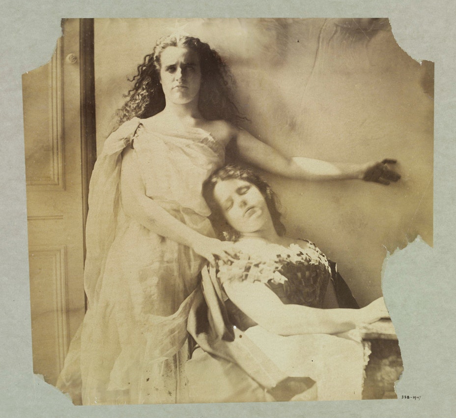 A sepia-toned photograph depicting two women in classical drapery. One is standing and looking directly at the camera with intense expression, and the other is reclining with her eyes closed, creating a dramatic tableau reminiscent of a scene from mythology or a historical painting.