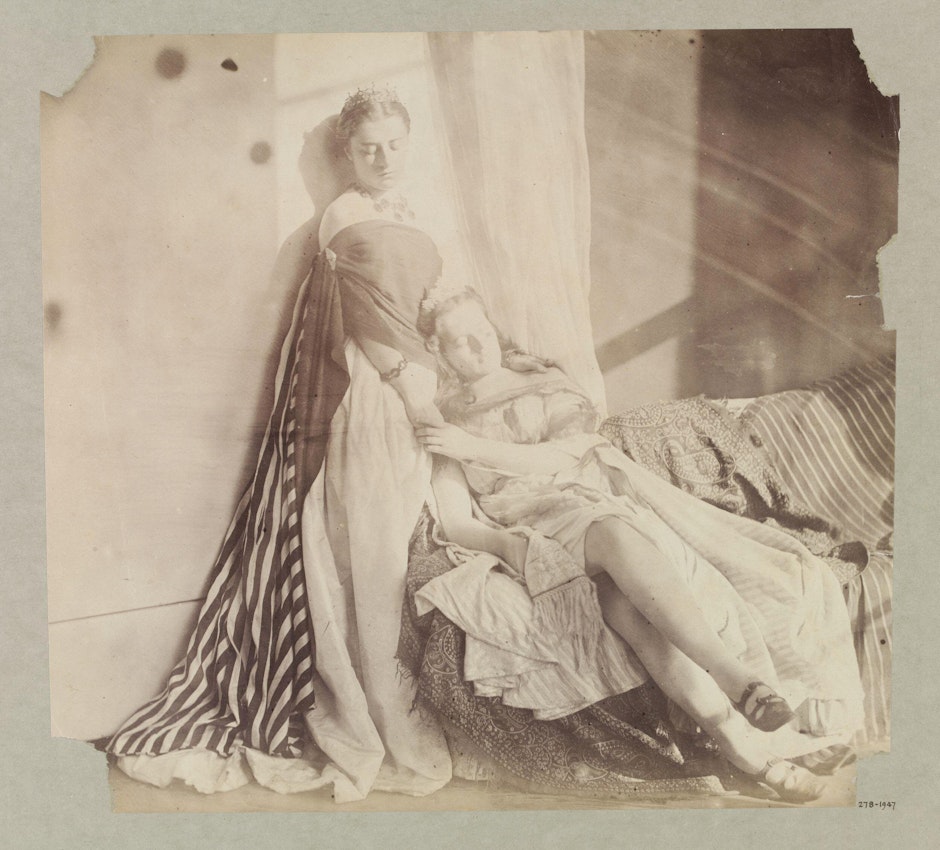 A sepia-toned photograph portraying two women in classical attire, one seated and the other lying in her lap, both in a relaxed, reclined pose with their eyes closed. The setting includes draped fabrics and a patterned throw.