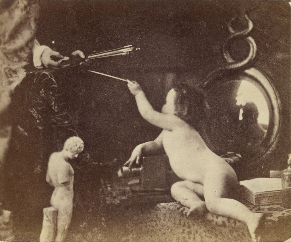 A photograph shows a nude infant handing handing a paintbrush to an adult standing out of frame whose hands are visible, holding a bundle of other paintbrushes. A mirror sits in the background behind the infant.