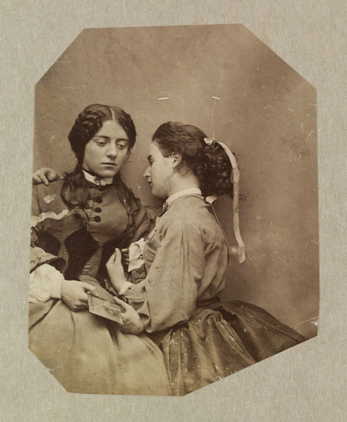 A sepia photograph of two women sitting closely, one holding a small photograph, with their faces turned towards each other.