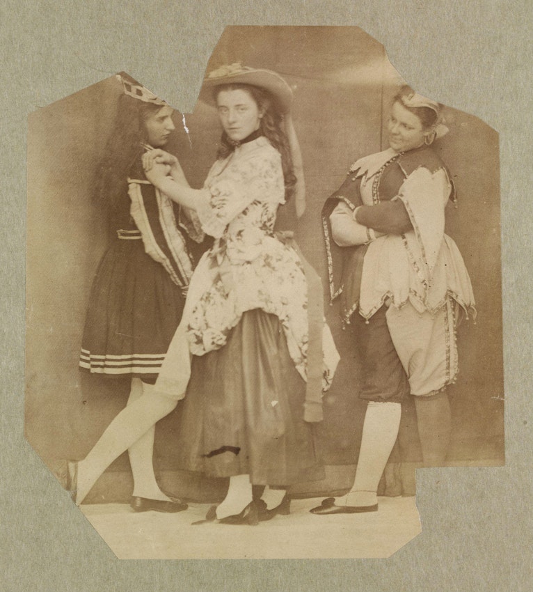 A sepia photograph of three women in costumes posed dramatically with one woman in the center looking directly at the camera, flanked by the others in profile.