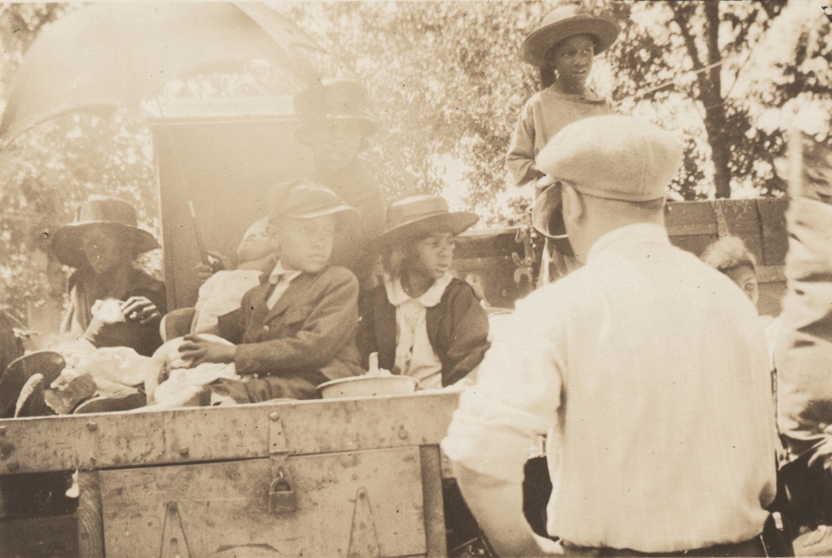 Black woman and children seated in wagon and watched over by white man