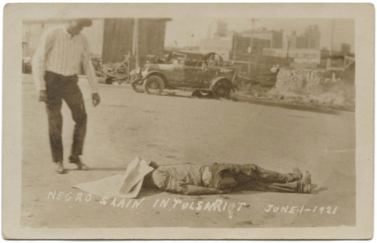 A white man stands over a dead person covered in the street